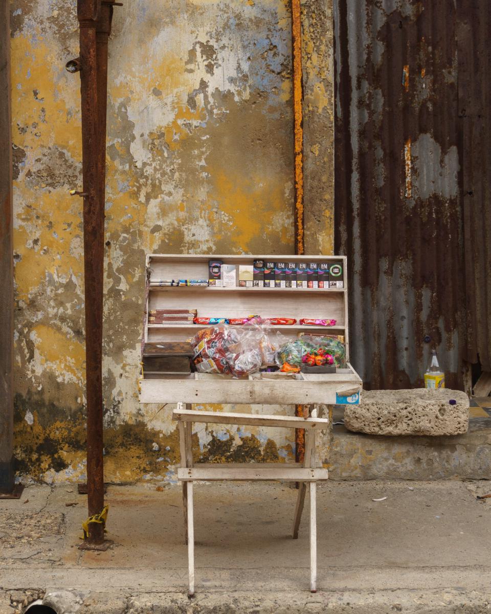 Estante de madera con venta de cigarrillos y otros objetos, muro descolorido de una casa antigua en proceso de remodelación